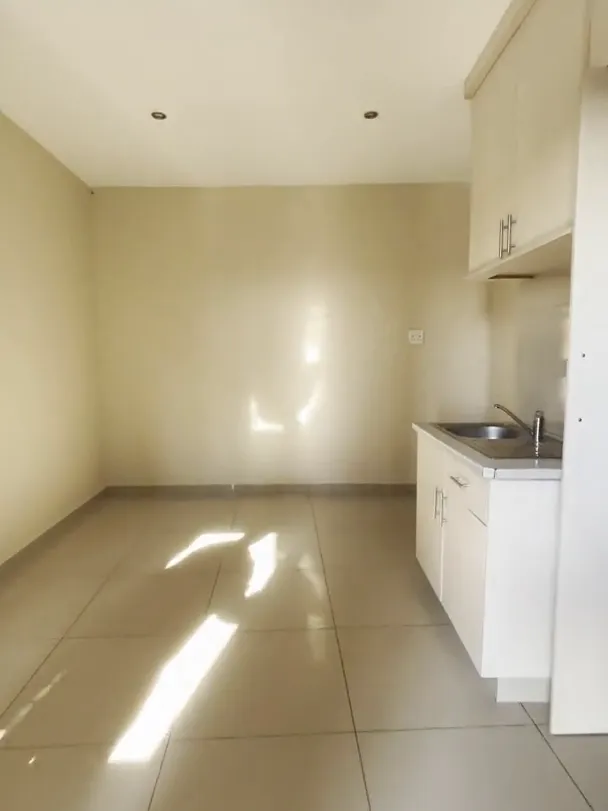 Empty room with a tiled floor, cream walls and a kitchenette with a sink and white cupboards along one side